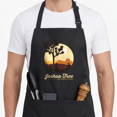 Joshua california tree apron - Circular desert sunset with tree silhouette and Joshua Tree National Park text — shown on a black cotton canvas apron against a white background