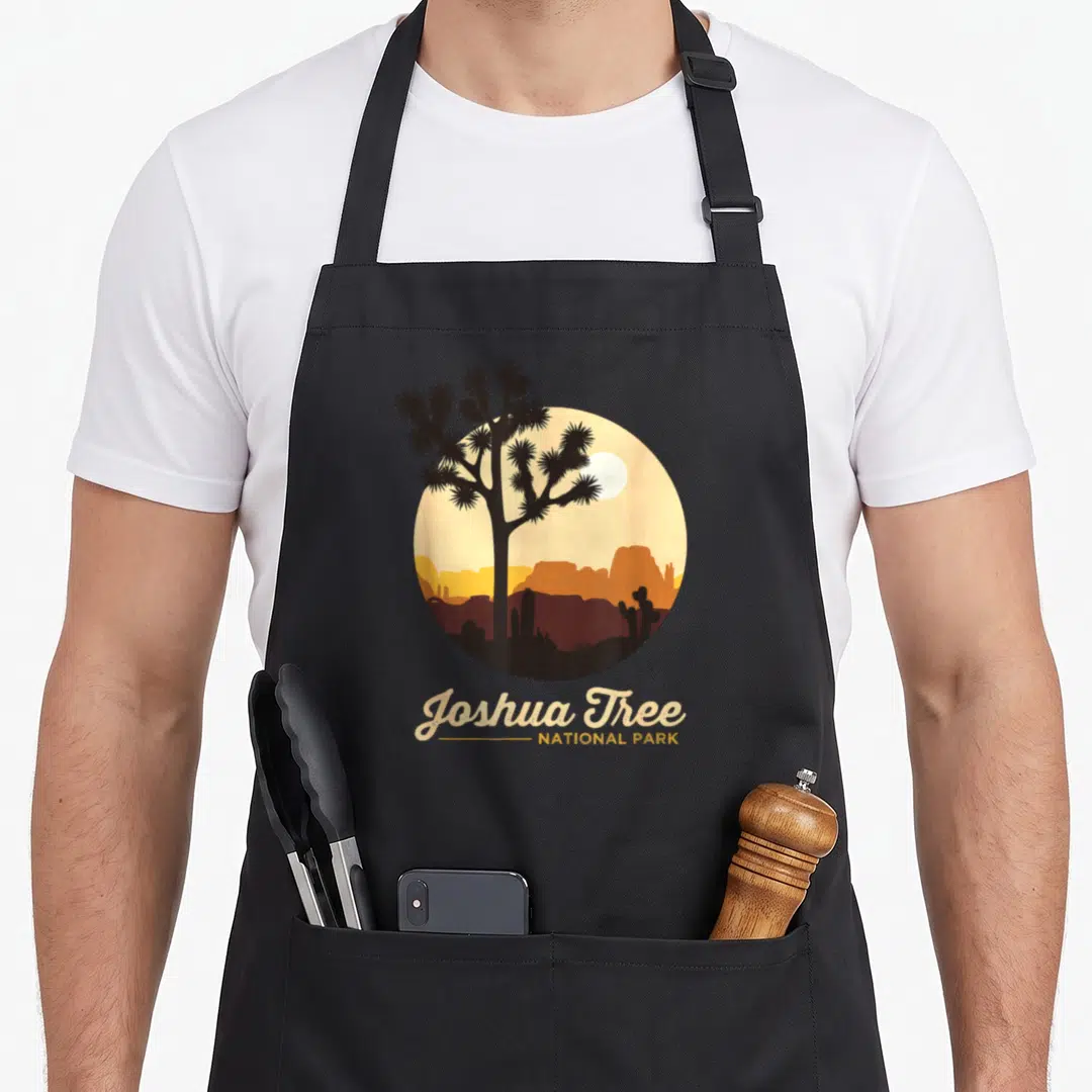 Joshua california tree apron - Circular desert sunset with tree silhouette and Joshua Tree National Park text — shown on a black cotton canvas apron against a white background