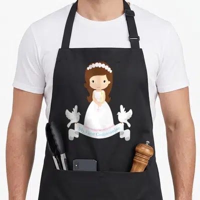 Rosary communion aprons - Cartoon girl in white dress holding candle and pink rosary with two doves and blue text banner — shown on a black cotton canvas apron against a white background
