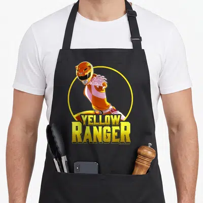 Portrait rangers apron - Yellow Ranger character in yellow circle with stacked YELLOW RANGER text — shown on a black cotton canvas apron against a white background