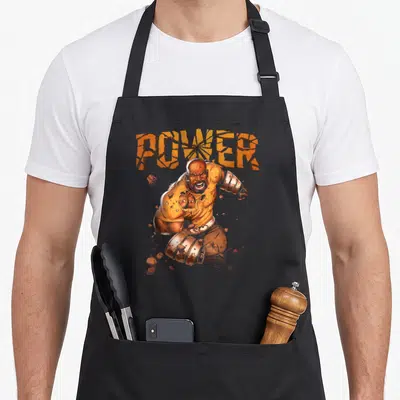 Luke hobby apron - Cracked orange POWER text above muscular man in orange brown tones surrounded by scattered rocks — shown on a black cotton canvas apron against a white background