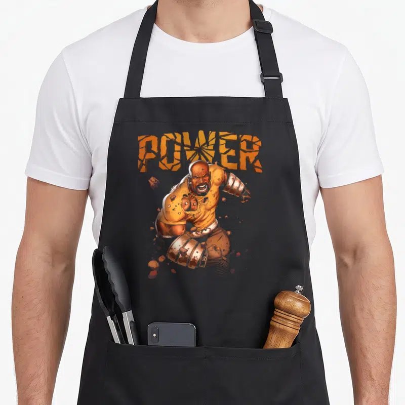Luke hobby apron - Cracked orange POWER text above muscular man in orange brown tones surrounded by scattered rocks — shown on a black cotton canvas apron against a white background