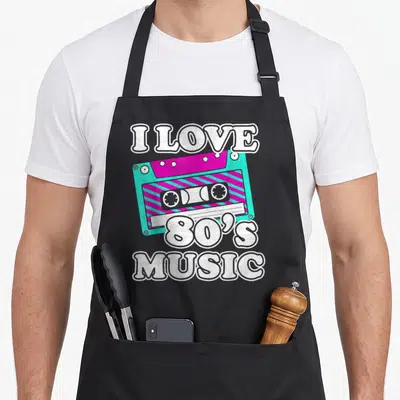 Music cassette 80s apron - Cartoon cassette tape in turquoise and magenta with white I LOVE 80'S MUSIC text — shown on a black cotton canvas apron against a white background