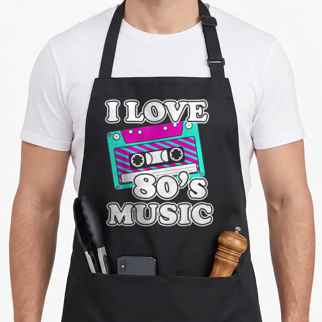 Music cassette 80s apron - Cartoon cassette tape in turquoise and magenta with white I LOVE 80'S MUSIC text — shown on a black cotton canvas apron against a white background