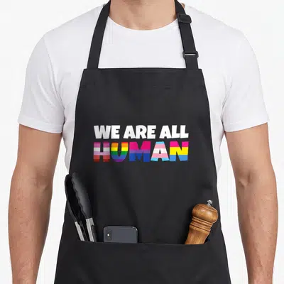 Flag rainbow apron - Stacked WE ARE ALL HUMAN text in rainbow and trans pride stripes — shown on a black cotton canvas apron against a white background