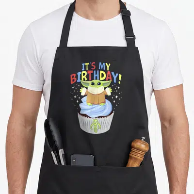 Funny birthday cupcake apron - Grogu standing on blue frosted cupcake with arched IT'S MY BIRTHDAY text and white stars — shown on a black cotton canvas apron against a white background
