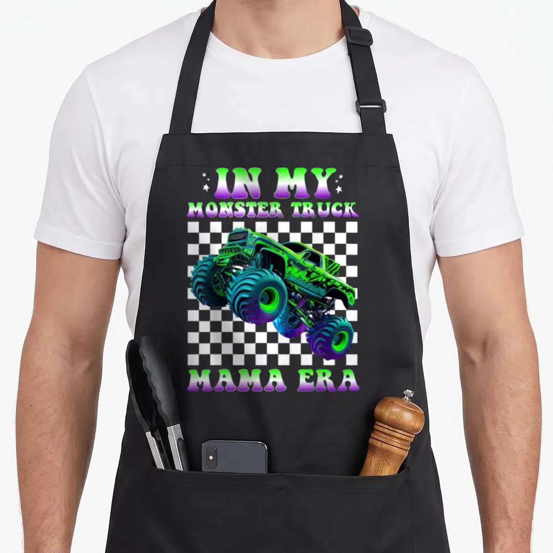 Groovy truck apron - Green purple blue monster truck on checkerboard with IN MY MONSTER TRUCK MAMA ERA text — shown on a black cotton canvas apron against a white background