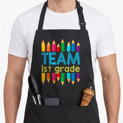 School 1st teacher apron - Colorful pencils surrounding blue TEAM and yellow 1st grade text on black bib — shown on a black cotton canvas apron against a white background