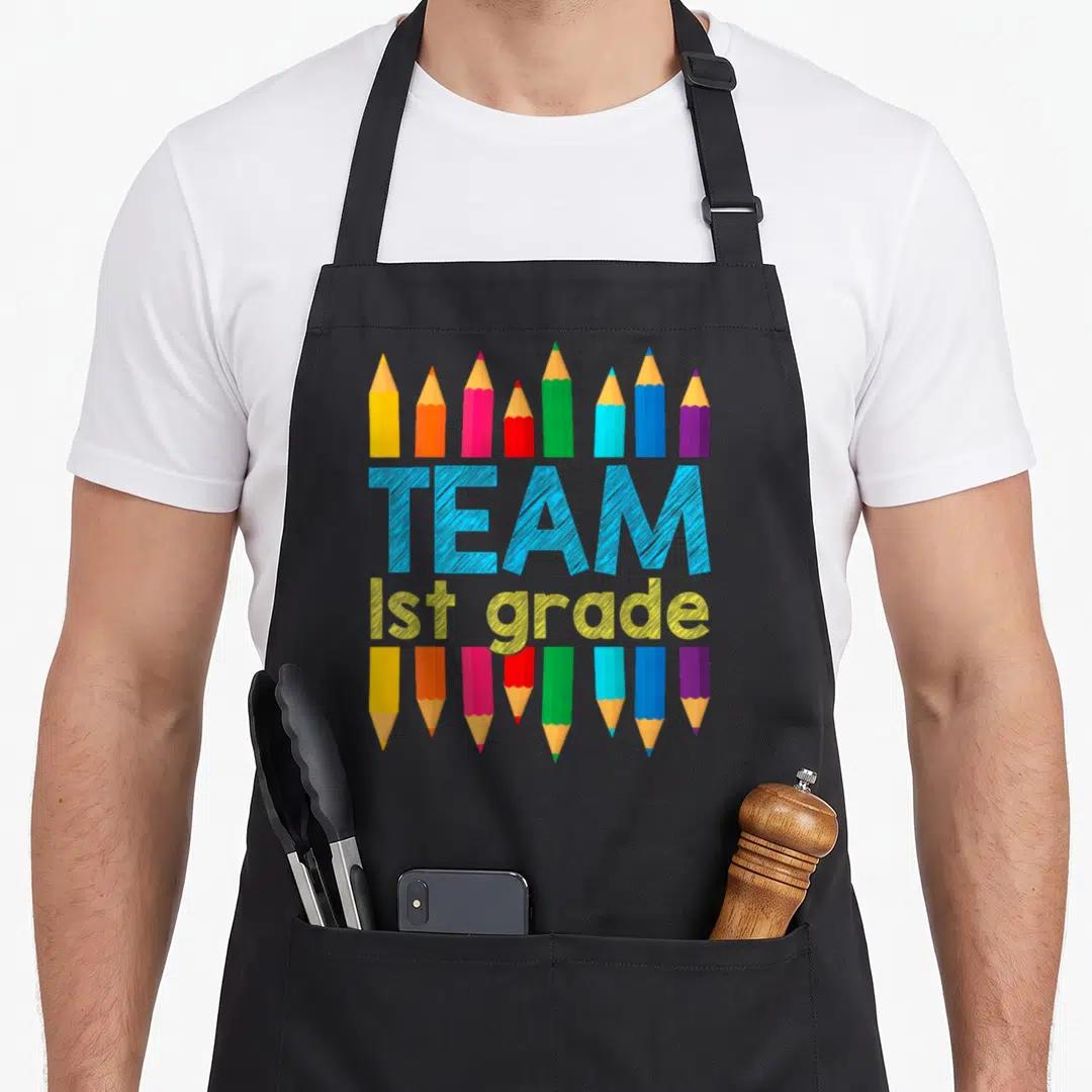 School 1st teacher apron - Colorful pencils surrounding blue TEAM and yellow 1st grade text on black bib — shown on a black cotton canvas apron against a white background