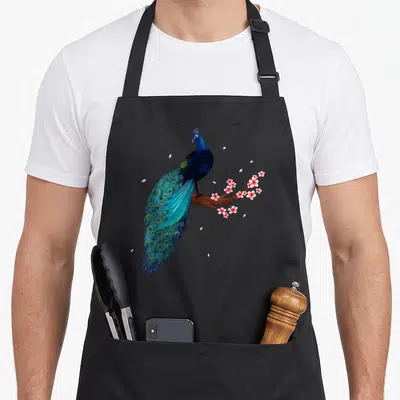 Beauty bird apron - Realistic peacock in blue and teal on brown branch with pink blossoms and scattered petals — shown on a black cotton canvas apron against a white background