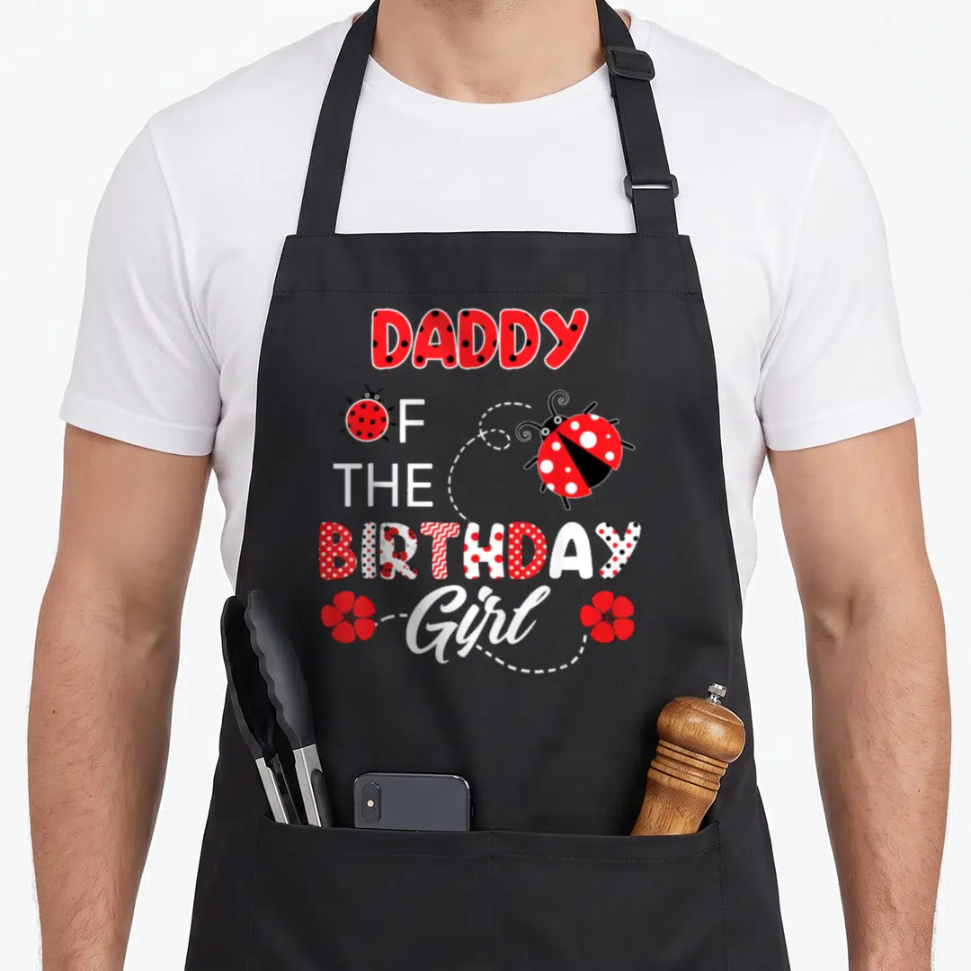Daddy ladybug apron - Stacked DADDY OF THE BIRTHDAY Girl text with red white black ladybugs and two red flowers linked — shown on a black cotton canvas apron against a white background