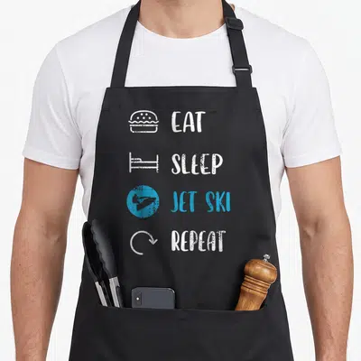 Rider jetski apron - Eat Sleep Jet Ski Repeat text with burger bed and jet ski icons in distressed white and cyan — shown on a black cotton canvas apron against a white background