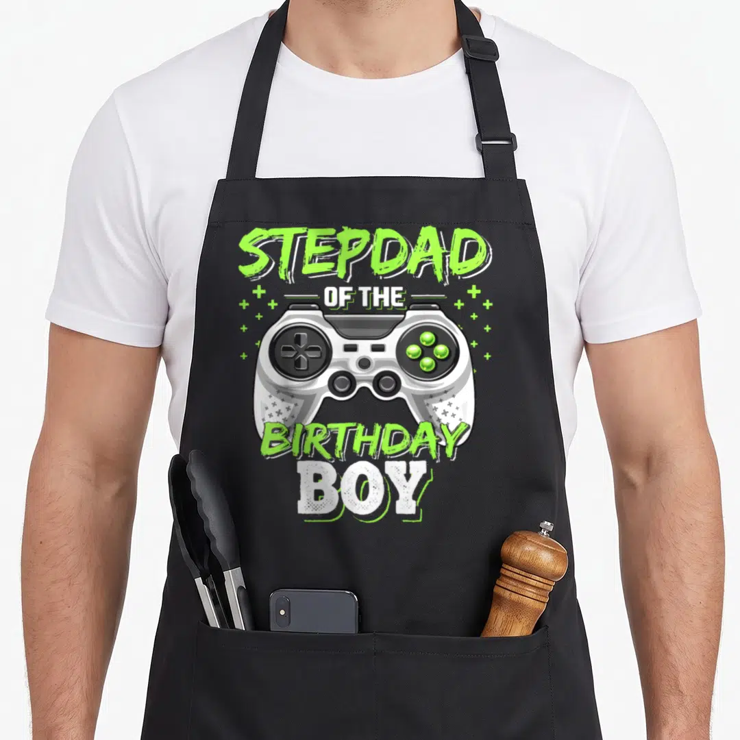 Stepdad video game apron - Grey game controller with lime green text STEPDAD OF THE BIRTHDAY BOY and plus signs — shown on a black cotton canvas apron against a white background
