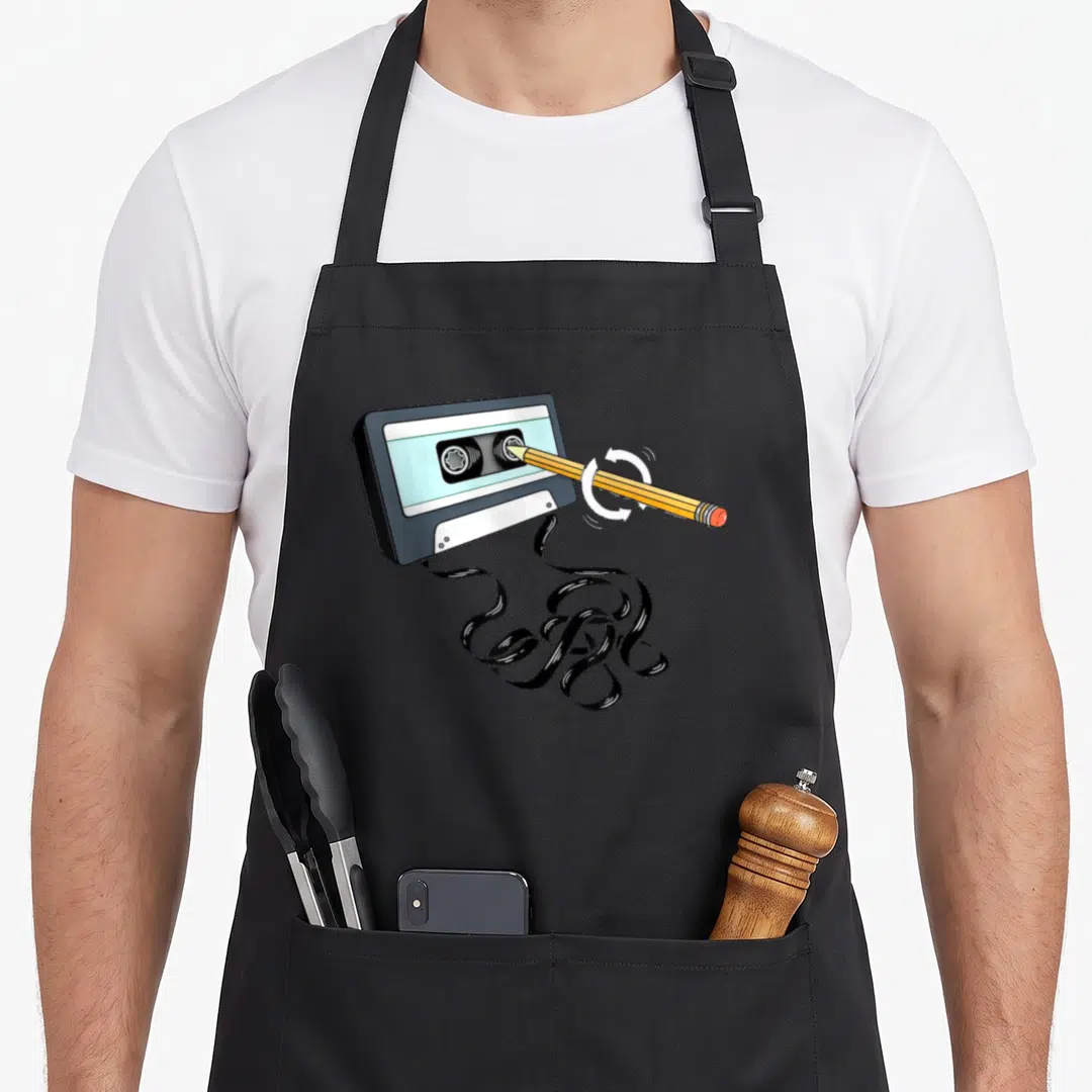 Pencil 80s apron - Cartoon cassette tape with pencil insertion, white arrows, and tangled black magnetic tape spill — shown on a black cotton canvas apron against a white background
