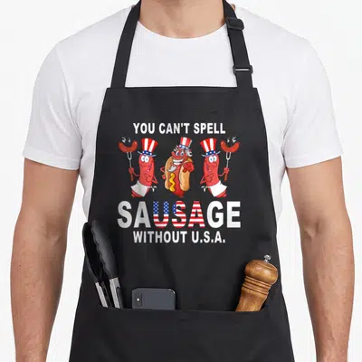 Spell sausage apron - Three cartoon sausages in patriotic hats with text YOU CAN'T SPELL SAUSAGE WITHOUT — shown on a black cotton canvas apron against a white background