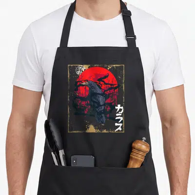 Raven moon gothic apron - Crow perched on branch before red sun with Japanese text and gold border — shown on a black cotton canvas apron against a white background
