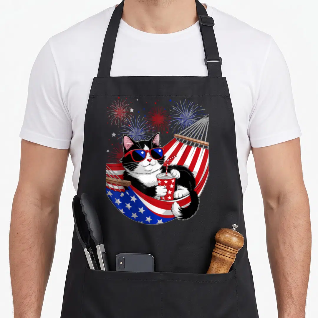 July tuxedo apron - Cartoon cat in sunglasses holding drink in red white blue striped hammock with fireworks — shown on a black cotton canvas apron against a white background