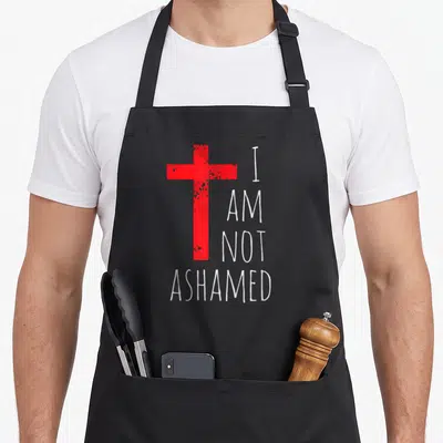 Ashamed red cross apron - Distressed red cross next to white text reading I AM NOT ASHAMED stacked vertically — shown on a black cotton canvas apron against a white background