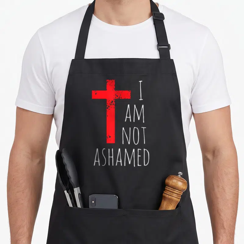 Ashamed red cross apron - Distressed red cross next to white text reading I AM NOT ASHAMED stacked vertically — shown on a black cotton canvas apron against a white background