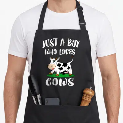 Cow farm aprons - Cartoon cow on green grass with text JUST A BOY WHO LOVES COWS — shown on a black cotton canvas apron against a white background