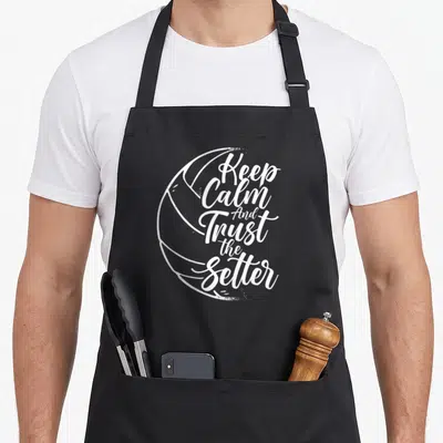 Trust setter apron - White stacked text reading Keep Calm And Trust the Setter inside a volleyball outline — shown on a black cotton canvas apron against a white background