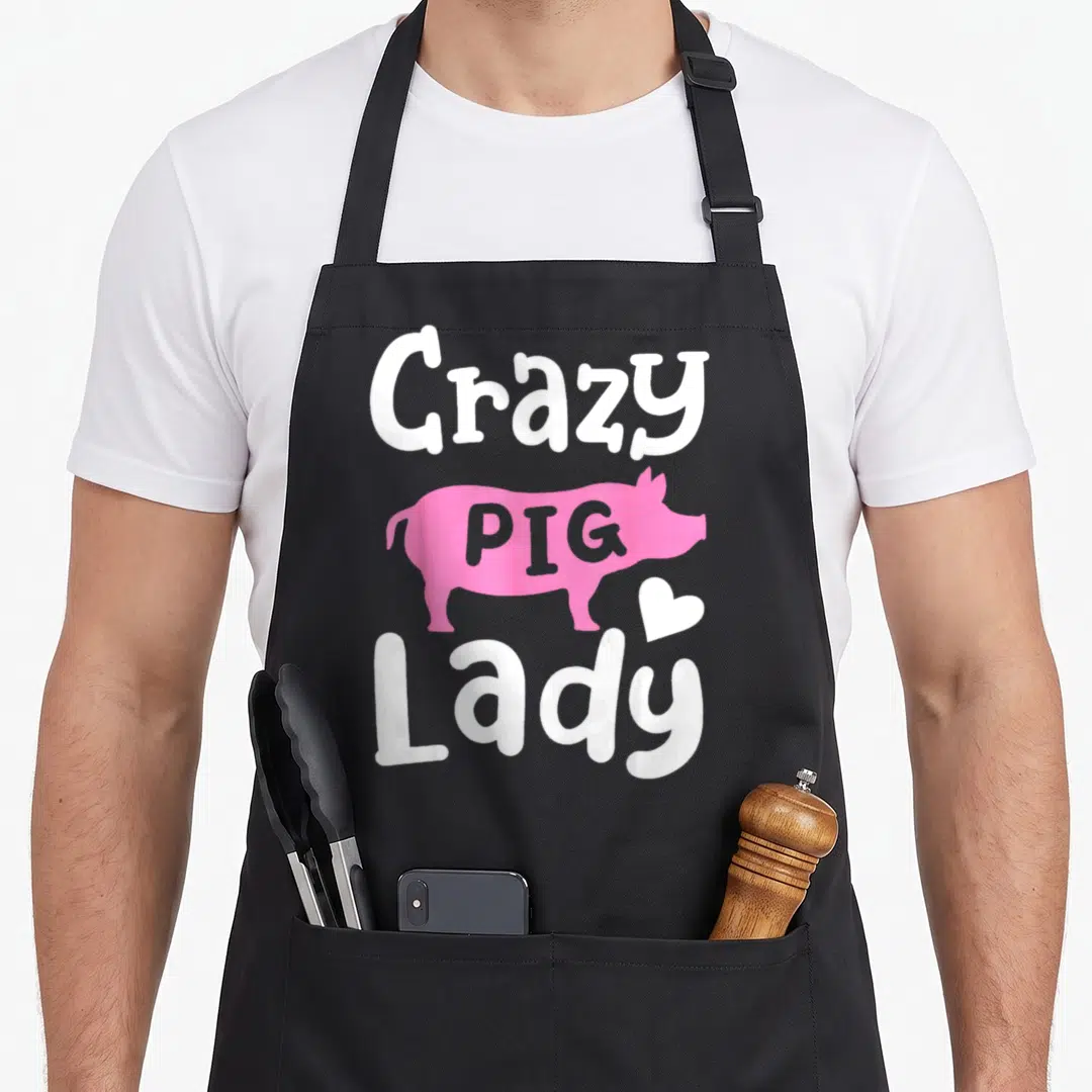 Funny crazy pig lady apron - Pink pig silhouette with Crazy PIG Lady text and white heart on black background — shown on a black cotton canvas apron against a white background