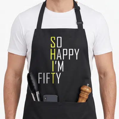 50th gag aprons - Stacked yellow and white text reading So Happy I'm Fifty — shown on a black cotton canvas apron against a white background
