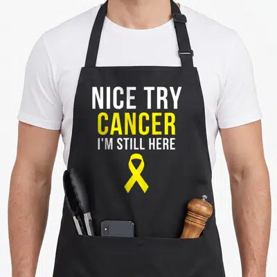 Try ribbon apron - NICE TRY CANCER I'M STILL HERE text with yellow awareness ribbon — shown on a black cotton canvas apron against a white background