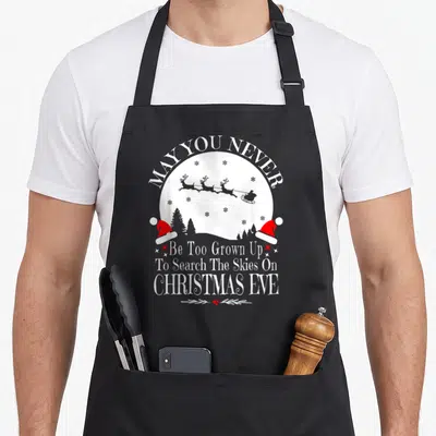 May christmas apron - Santa sleigh in white moon with red hats and text May You Never Be Too Grown Up — shown on a black cotton canvas apron against a white background