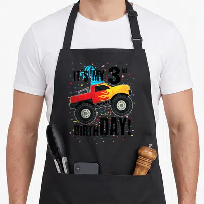 3rd birthday monster truck apron - Cartoon monster truck with party hat and IT'S MY 3RD BIRTHDAY text amidst confetti — shown on a black cotton canvas apron against a white background