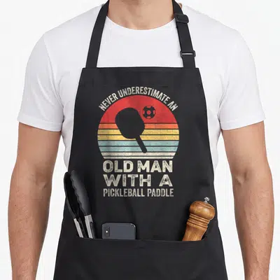 Vintage underestimate pickleball apron - Retro striped sun with pickleball paddle silhouette and white vintage lettering — shown on a black cotton canvas apron against a white background