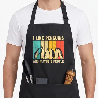 Penguin aprons for cooking - Six penguins standing before beige teal yellow orange red vertical stripes with text — shown on a black cotton canvas apron against a white background