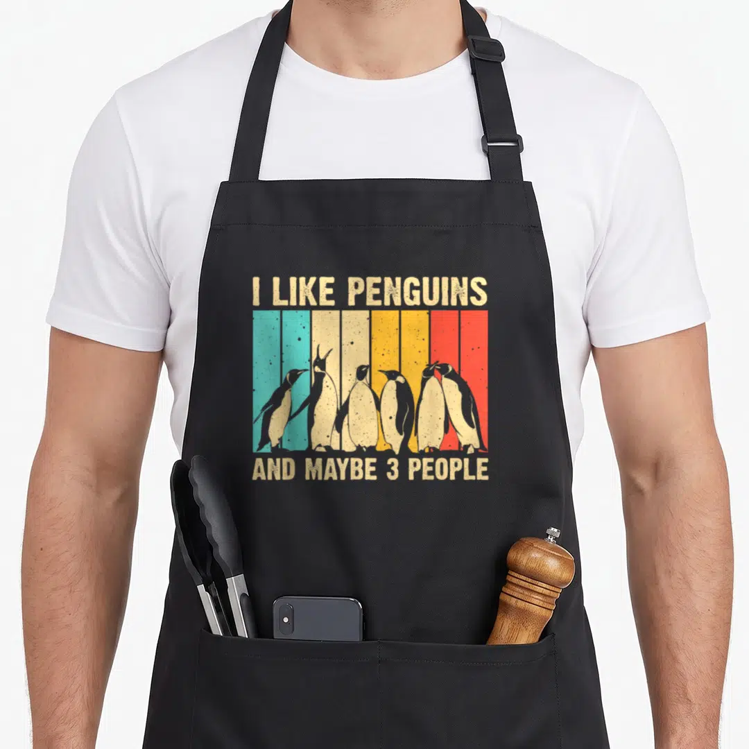 Penguin aprons for cooking - Six penguins standing before beige teal yellow orange red vertical stripes with text — shown on a black cotton canvas apron against a white background