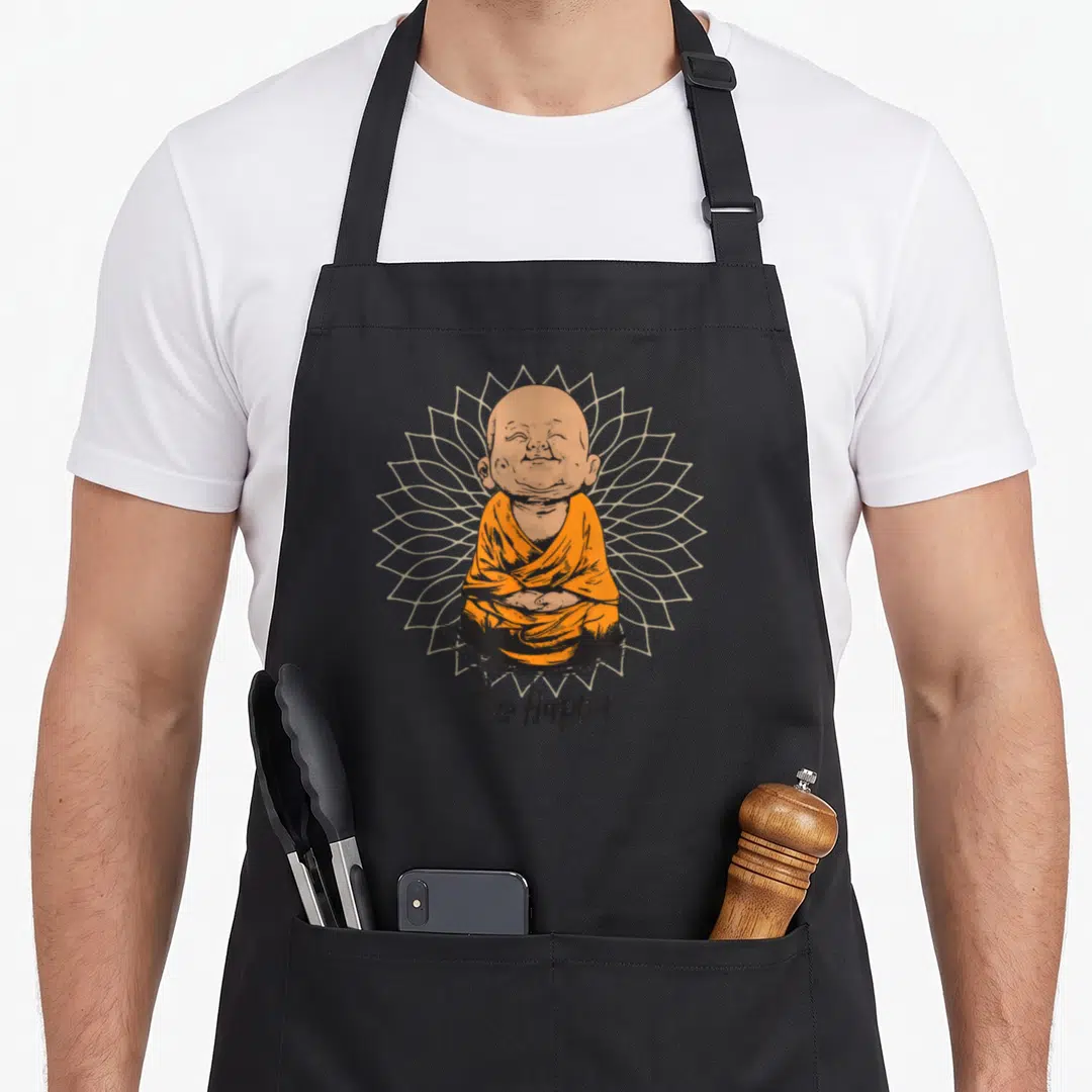 Zen meditation buddhist apron - Smiling cartoon monk in orange robes sitting before beige mandala with Be Happy text — shown on a black cotton canvas apron against a white background