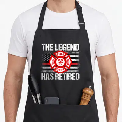 Legend firefighter cooking apron - Distressed flag Maltese cross with ladder axes hydrant and THE LEGEND HAS RETIRED — shown on a black cotton canvas apron against a white background