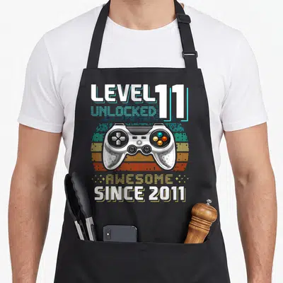 Age video game apron - White controller over pixel sun with Level 11 Unlocked text — shown on a black cotton canvas apron against a white background