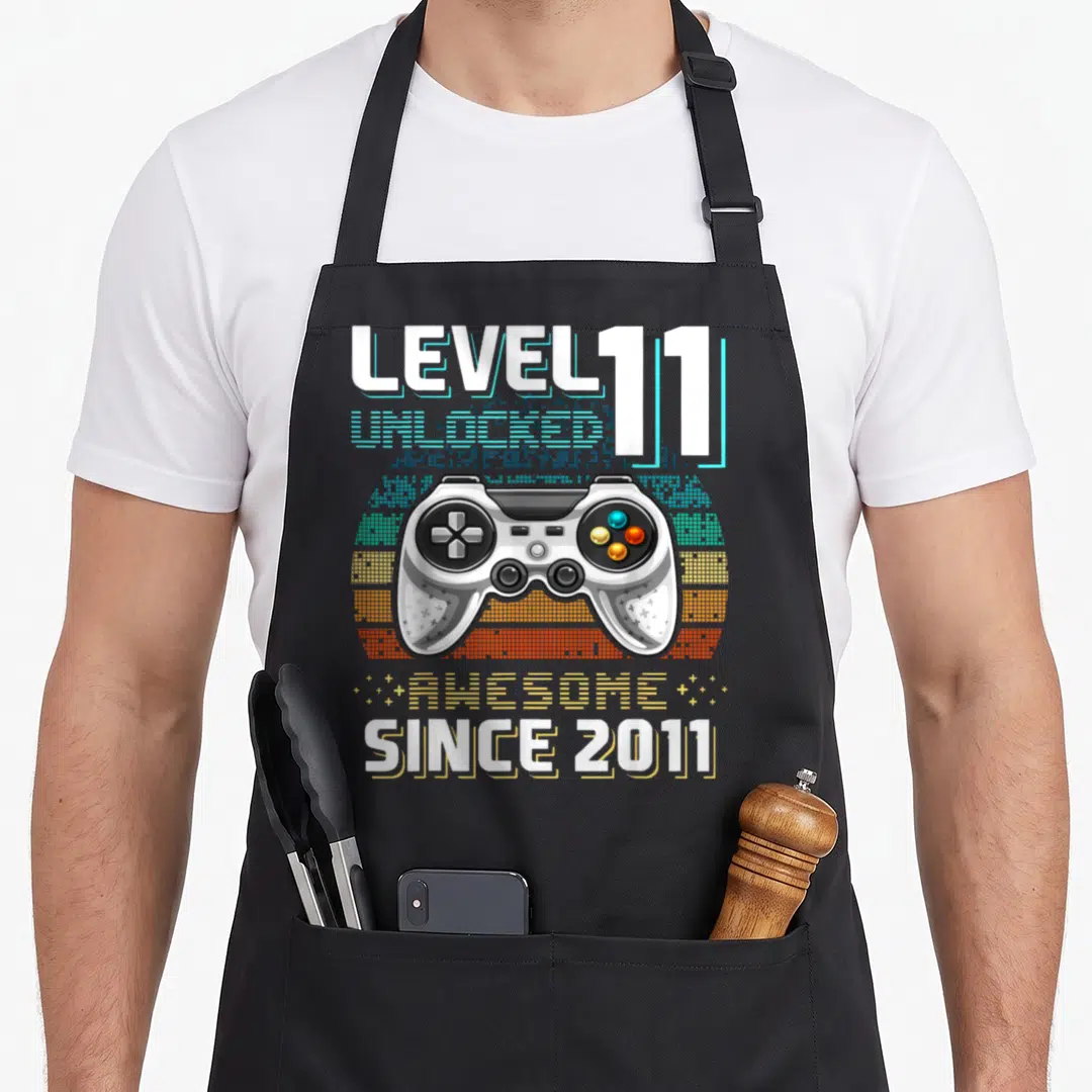 Age video game apron - White controller over pixel sun with Level 11 Unlocked text — shown on a black cotton canvas apron against a white background