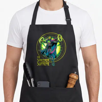 Sorcerer doctor apron - Doctor Strange floating in yellow circle with green rings and stacked yellow text reading — shown on a black cotton canvas apron against a white background