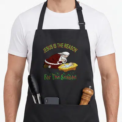 Kneeling santa apron - Cartoon Santa kneeling by manger under JESUS IS THE REASON For The Season text — shown on a black cotton canvas apron against a white background
