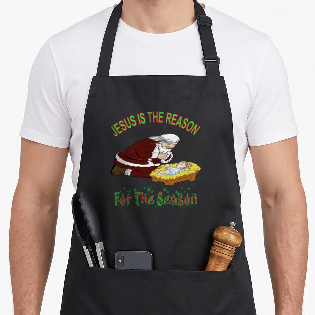 Kneeling santa apron - Cartoon Santa kneeling by manger under JESUS IS THE REASON For The Season text — shown on a black cotton canvas apron against a white background