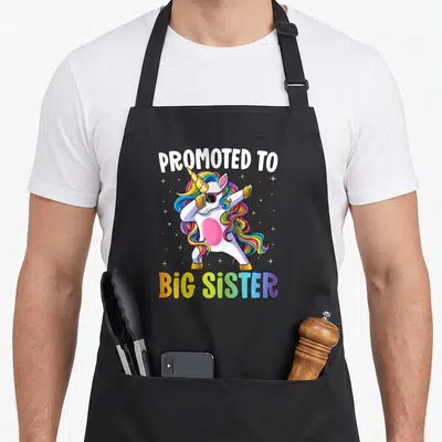 Promoted unicorn apron - Dabbing unicorn with rainbow mane and PROMOTED TO BIG SISTER text surrounded by stars — shown on a black cotton canvas apron against a white background