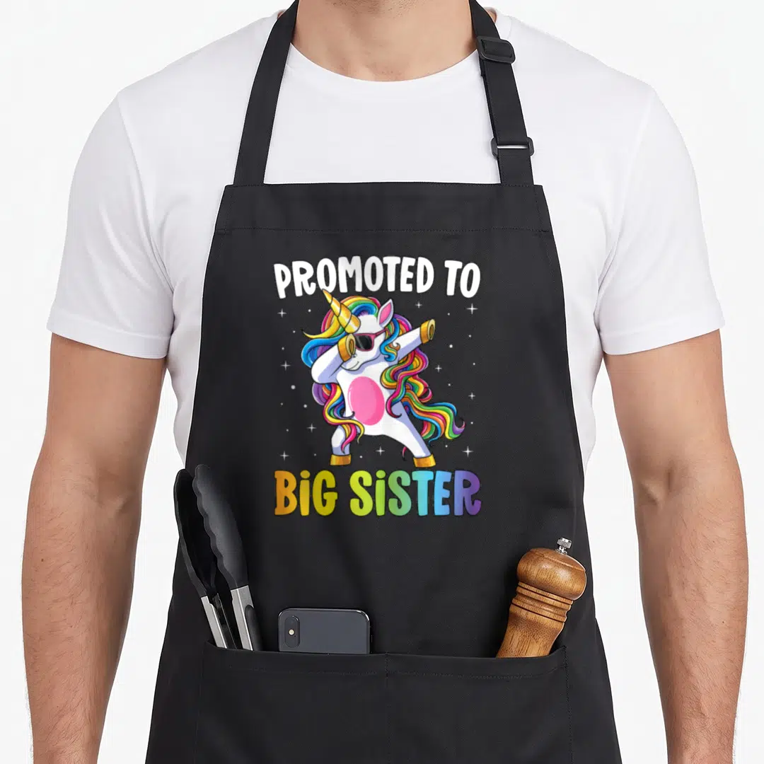 Promoted unicorn apron - Dabbing unicorn with rainbow mane and PROMOTED TO BIG SISTER text surrounded by stars — shown on a black cotton canvas apron against a white background