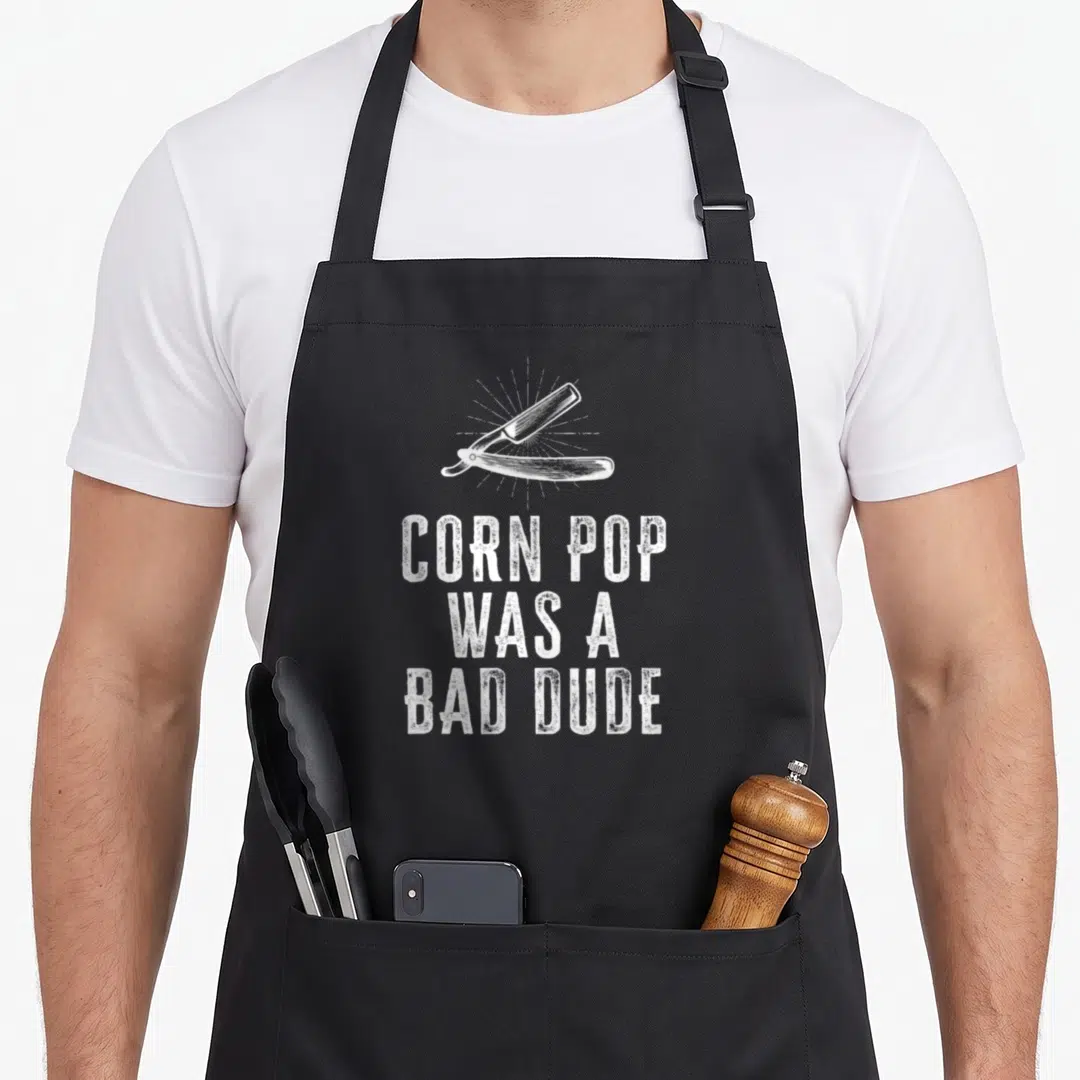 Corn dude apron - White straight razor over sunburst with stacked text CORN POP WAS A BAD DUDE — shown on a black cotton canvas apron against a white background