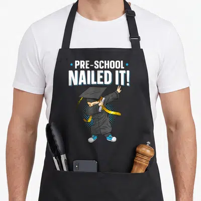 Dabbing preschool apron - Text PRE-SCHOOL NAILED IT! above a boy in a graduation cap dabbing with a diploma scroll — shown on a black cotton canvas apron against a white background