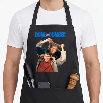 Dumb key apron - DUMB AND DUMBER text above two men, one pulling other's hair, blue grey red photo style — shown on a black cotton canvas apron against a white background