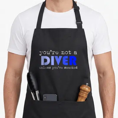 Water athletic lettering apron - Stacked text reading you're not a diver unless you've smacked in blue and white — shown on a black cotton canvas apron against a white background