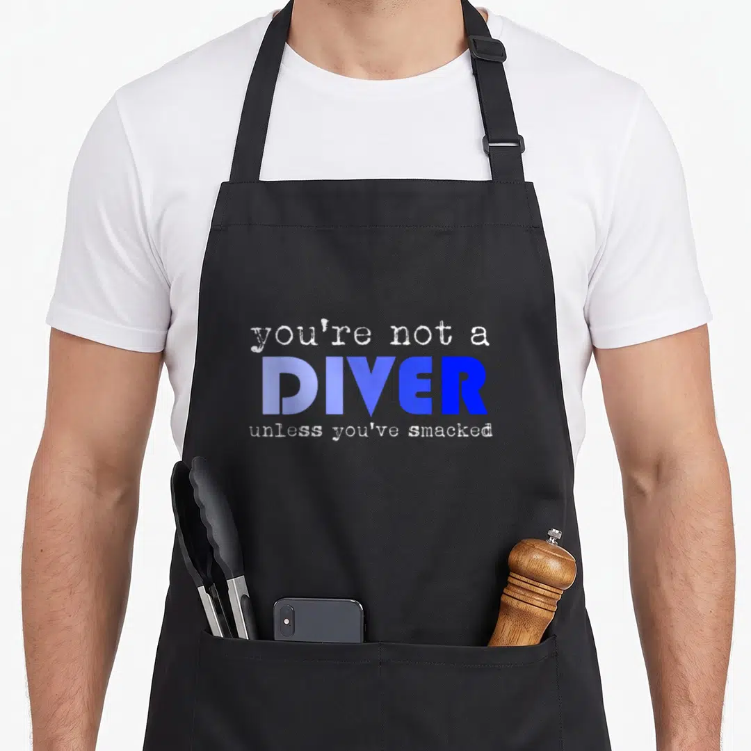 Water athletic lettering apron - Stacked text reading you're not a diver unless you've smacked in blue and white — shown on a black cotton canvas apron against a white background