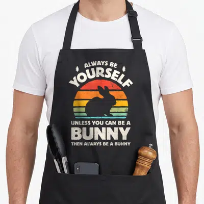 Alway bunny apron - Retro striped sun with white bunny silhouette and text Always Be Yourself Unless You Can Be A Bunny — shown on a black cotton canvas apron against a white background