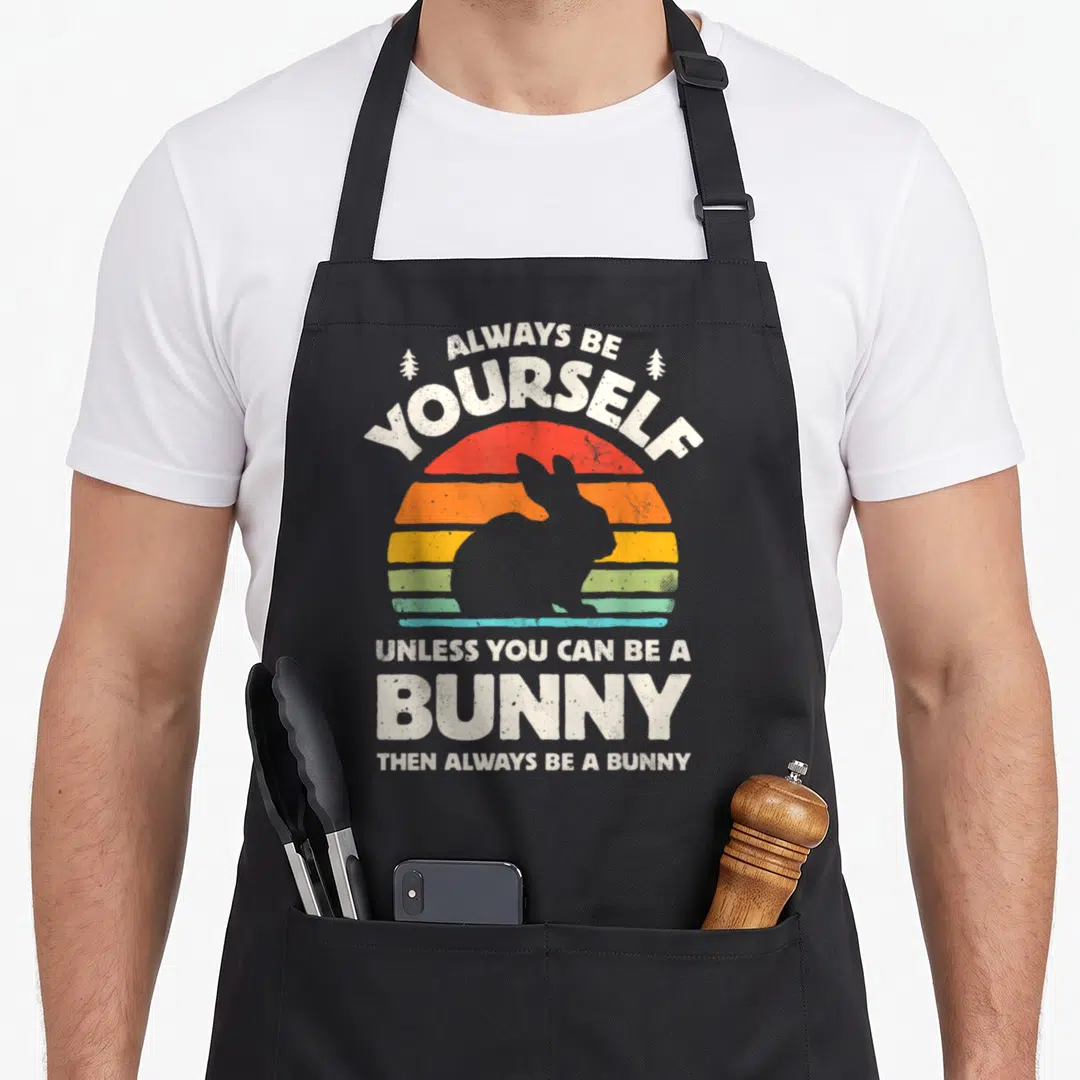 Alway bunny apron - Retro striped sun with white bunny silhouette and text Always Be Yourself Unless You Can Be A Bunny — shown on a black cotton canvas apron against a white background