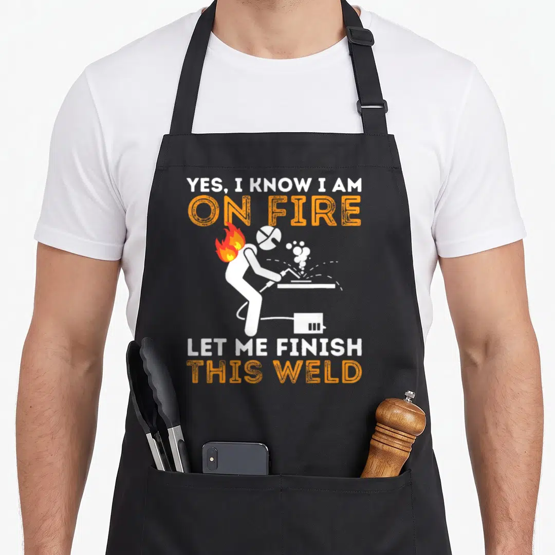 Fire metal worker apron - Cartoon welder with flames on helmet and text YES I KNOW I AM ON FIRE LET ME FINISH THIS WELD — shown on a black cotton canvas apron against a white background